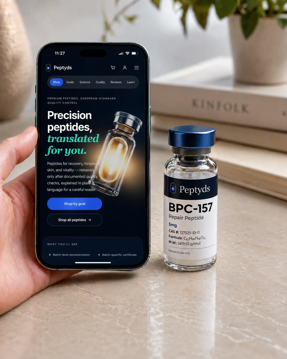 Hand holding a phone showing the Peptyds homepage hero next to a real BPC-157 5 mg vial on a marble counter