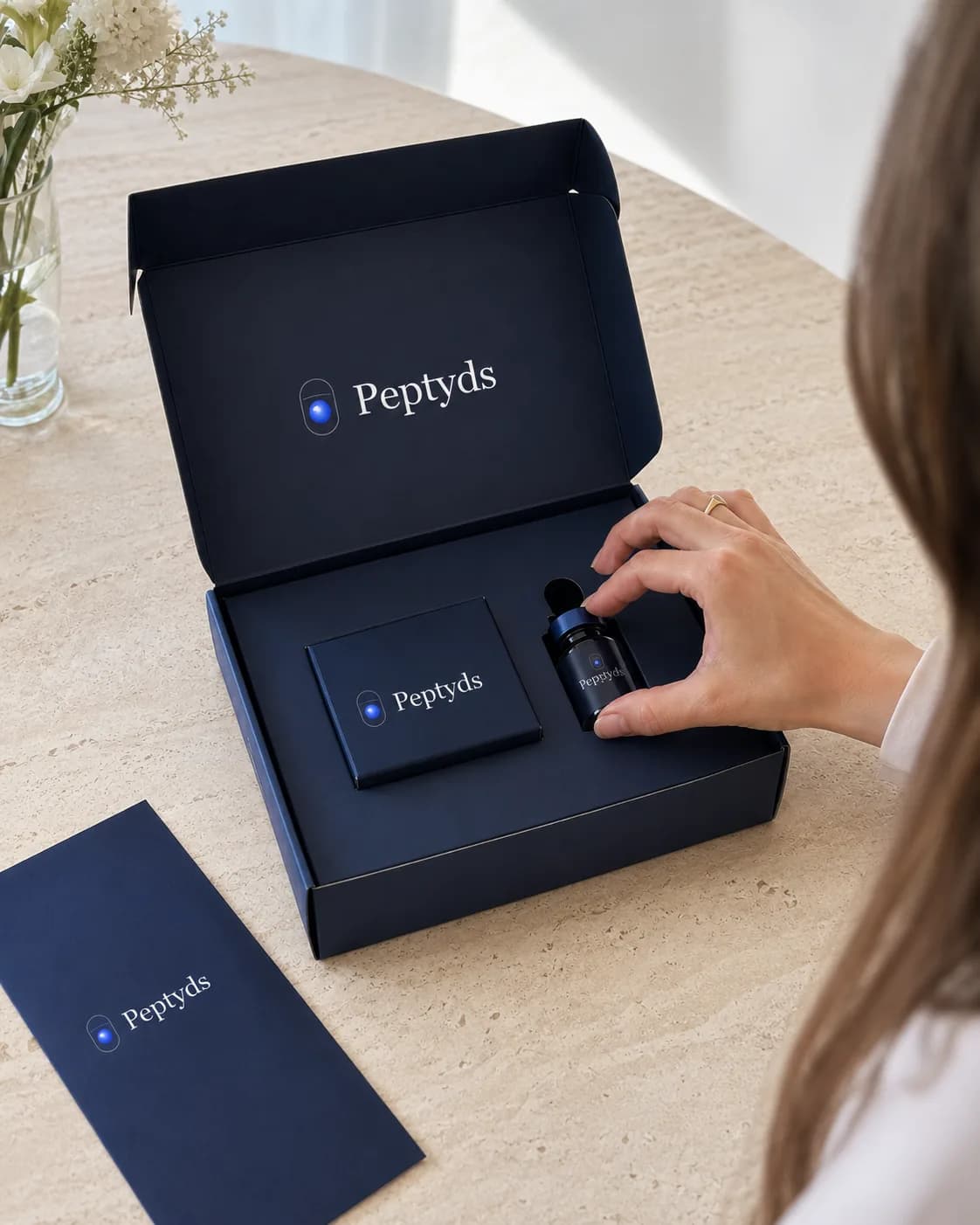 Unboxing eines dunkelblauen Peptyds-Pakets auf hellem Marmor aus der Vogelperspektive — Peptid-Vial, Umkarton, Umschlag und weiße Blumen im Weichzeichner